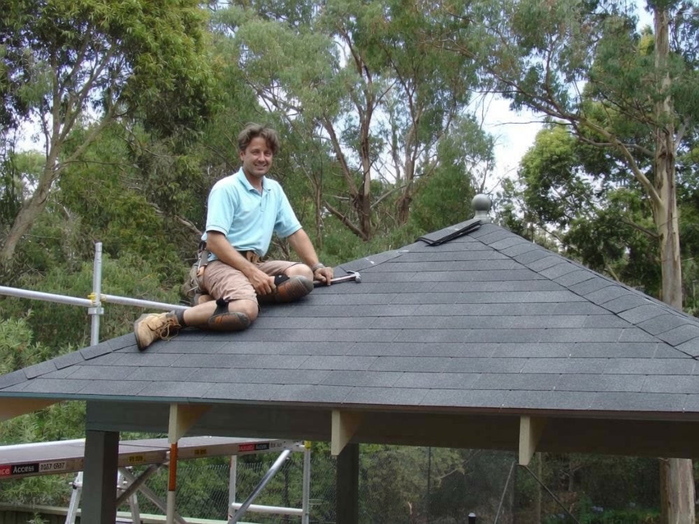 Outdoor Gazebos Australia 22 Man sitting on a dual black asphalt shingle roof while installing shingles on a timber gazebo kit, diy roofing project